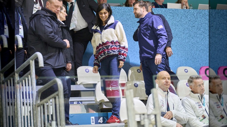 Usha Vance and JD Vance speak to security guards at the 2026 Winter Olympics. Usha is wearing red sneakers with a white stripe, navy blue pants, and a white wool jacket.