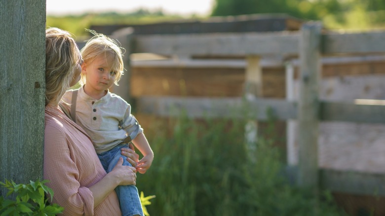 A woman leaning on an outdoor fence holds her toddler.