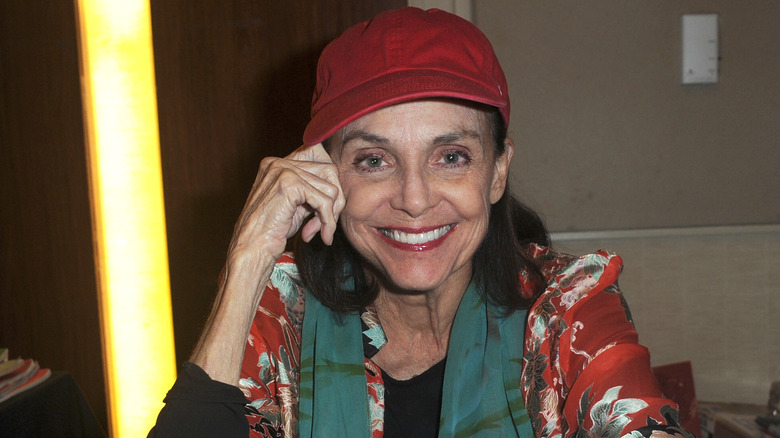 Valerie Harper sitting  at a table smiling and wearing a red hat.