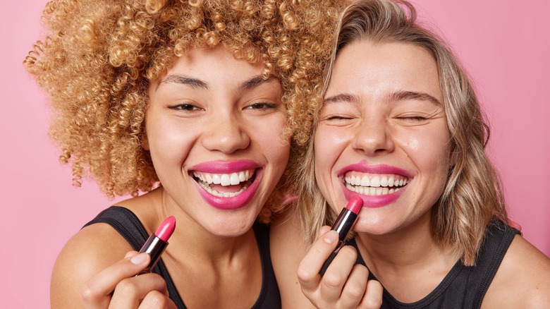 Two women smile applying lipstick