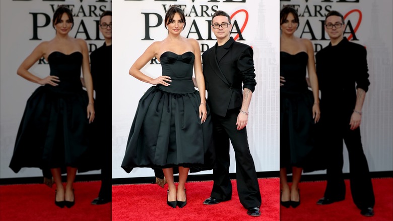 Paige DeSorbo poses with Christian Siriano. DeSorbo is a young woman with fair skin and dark hair. She is wearing a black sleeveless dress with a drop-waist A-line skirt. Christian Siriano is a man with pale skin and short dark hair. He is wearing a black suit with sleeves pushed up his arms.