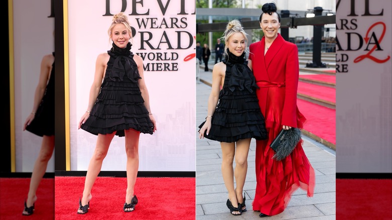 Tara Lipinski and Johnny Weir at the DWP2 premiere. Tara is a woman with fair skin and blonde hair pulled up into a messy top knot. She is wearing a short black ruffled babydoll dress. Johnny is a man with pale skin and dark hair also pulled up into a top knot. He is wearing a bright red blazer style coat with matching pants and a fringed black clutch bag.