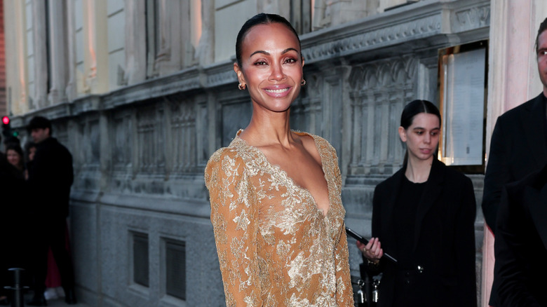 Zoe Saldaña posing at an event wearing a beige dress with gold embellishments.