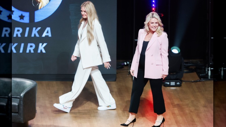 Full image of Kirk and Leavitt at the Turning Point USA event. Kirk is wearing a baggy white pantsuit and sneakers. Leavitt is wearing a light pink blazer