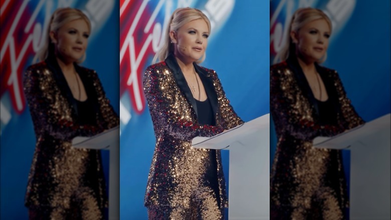Erika Kirk wears a gold sequin suit with a black lapel while speaking at the podium of a TPUSA event. Her blonde hair is tied back in a high ponytail