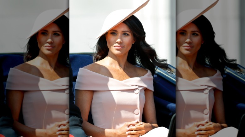 The full image of Markle's 2018 Trooping The Colours look. She is sitting in a carriage wearing a pale pink off the shoulder dress without sleeves.