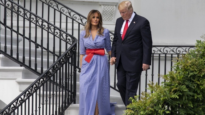 Melania and Donald Trump walk down the White House stairs (zoomed out). Melania is wearing a white and blue striped button down dress with a large red bow belt.