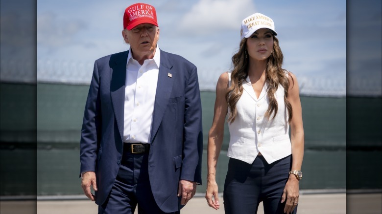 Donald Trump stands next to Kristi Noem in front of a dark green fence